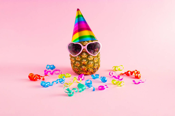 Pineapple with hat, glasses and colorul party streamers on pink background. Minimal party and celebration concept. - Foto, imagen