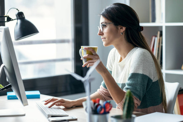 Снимок сконцентрированной молодой деловой женщины, работающей за компьютером, когда она пила кофе в современном офисе стартапа. - Фото, изображение
