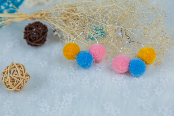 Σκουλαρίκια pompon χρωματιστά σε σχήμα τριών χνουδωτών σφαιρών. Σκουλαρίκια είναι δίπλα σε προϊόντα μπαστούνι, δείγματα λουλουδιών, και άλλα στολίδια - Φωτογραφία, εικόνα