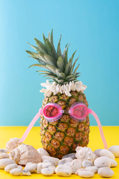 Piña en gafas de baño de color rosa con guijarros de mar y conchas marinas de cerca sobre un fondo amarillo-azul, concepto de verano - Foto, imagen