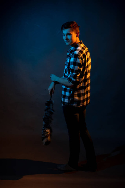A guy in a plaid shirt with a dust brush on a dark background illuminated by blue and yellow light - Foto, afbeelding