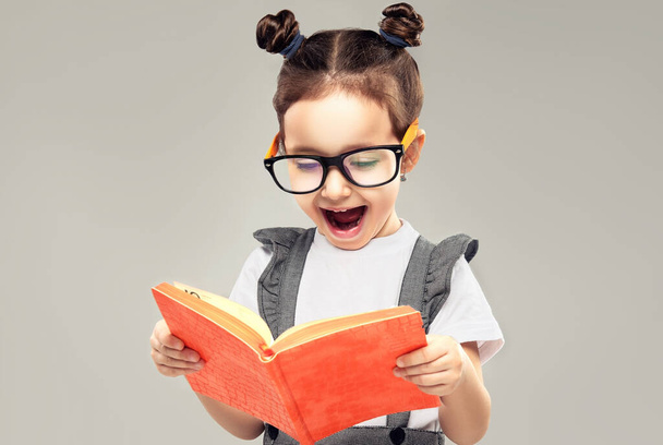 Child girl reads a book in surprise. A kid in glasses looks in amazement at an orange notebook. Children's products , clothing and accessories . Expressive facial emotions - Photo, Image