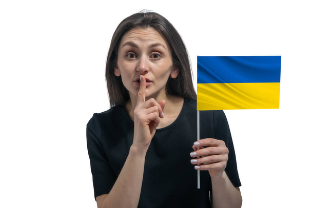 Heureuse jeune femme blanche tenant le drapeau de l'Ukraine et tenant un doigt sur ses lèvres isolées sur un fond blanc. - Photo, image