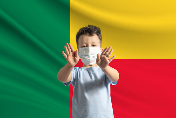 Mały biały chłopiec w masce ochronnej na tle flagi Beninu. Robi znak stopu rękami, zostaje w domu Benin. - Zdjęcie, obraz