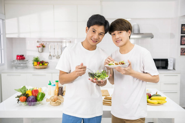 Χαμογελαστό ζευγάρι Ασιατών γκέι μαγειρεύουν σαλάτα και σάντουιτς για πρωινό. Χαρούμενη νεανική ΛΟΑΤ ζωή στο σπίτι. Ομοφυλόφιλη οικογένεια του ίδιου φύλου με υγιεινό τρόπο ζωής τρώνε. - Φωτογραφία, εικόνα