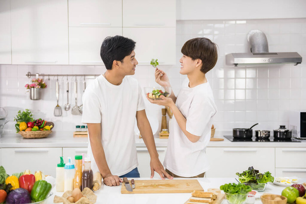 Cute Asian male gay feed vegetable salad to his partner after cooking for breakfast at kitchen. Happy young LGBT life at home. Homosexual same sex family with healthy eat lifestyle. - Photo, Image