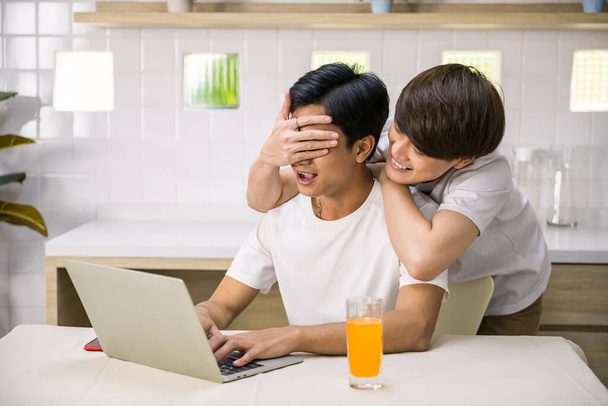 Leuke Aziatische man homo hand sluit zijn partner ogen om te voorkomen dat hij te werken op de computer in het weekend. Gelukkig LGBT koppel leven thuis. Homoseksuele familie van hetzelfde geslacht. - Foto, afbeelding