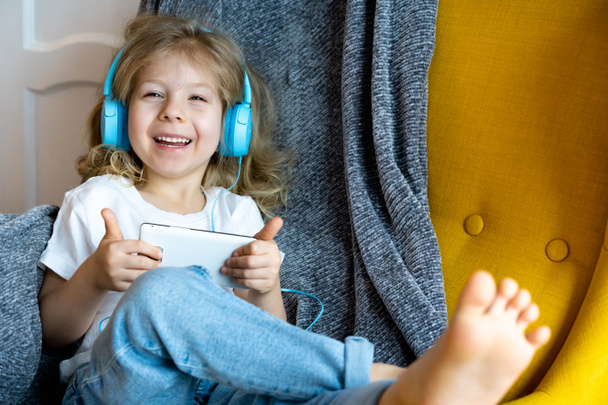 A little happy blonde at home in a chair listening to music with headphones and playing in a smartphone, the girl smiles. - Zdjęcie, obraz