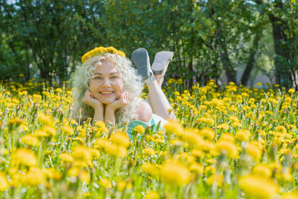 Byl jasný slunečný den. Mladá šťastná blondýnka leží na trávníku ze žlutých pampelišek a usmívá se. Výborná jarní slavnostní nálada. Venkovní rekreace. - Fotografie, Obrázek