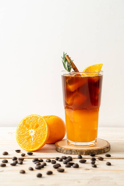 Бокал черного кофе со льдом и слой апельсинового и лимонного сока, украшенный розмарином и корицей на деревянном фоне - Фото, изображение