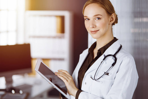 Cheerful smiling female doctor using clipboard in clinic. Portrait of friendly physician woman at work. Medical service in hospital. Medicine concept - Photo, image