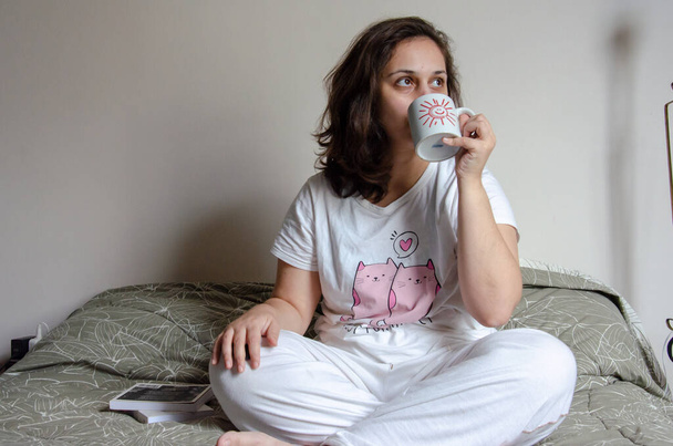 A young Brazilian female drinking coffee and relaxing on her bed - Zdjęcie, obraz
