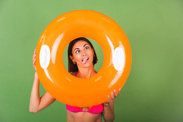 Summer fit sporty woman in pink bikini and bright orange inflatable ring round on green background, happy playful with funny grimaces - Fotografie, Obrázek