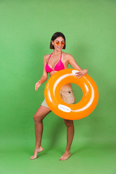 Summer fit sporty woman in pink bikini and bright orange inflatable ring round and sunglasses on green background, happy cheerful excited joyful positive - Photo, Image