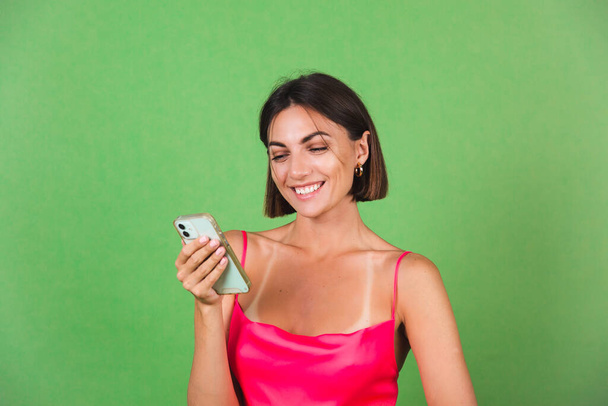Stylish woman in pink silk dress isolated on green background happy with a smile look on mobile phone screen, read message news - Φωτογραφία, εικόνα