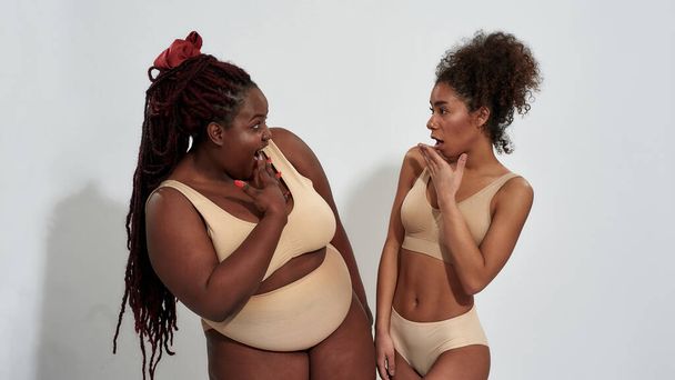 Plus size and slim african american women in underwear gossiping, looking emotional at each other, standing isolated over gray background - Fotografie, Obrázek