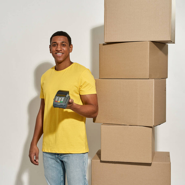 Smiling male mover in yellow t shirt holding terminal for payment with credit card, standing near a stack of cardboard boxes while doing home relocation - Fotoğraf, Görsel