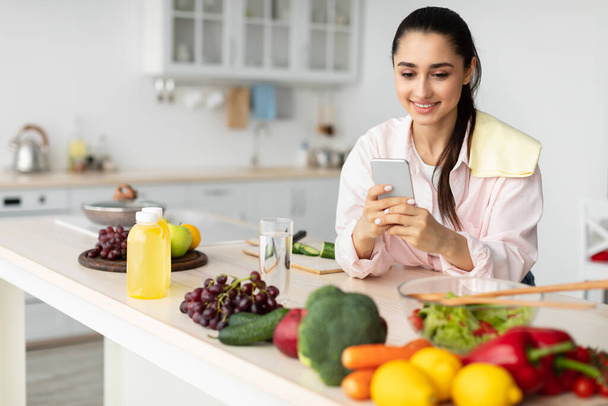 Портрет улыбающейся девушки, использующей мобильный телефон на кухне - Фото, изображение