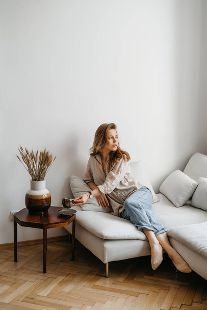 Μια νεαρή ξανθιά γυναίκα σε casual άνετα ρούχα κάθεται σε ένα λευκό καναπέ στο σπίτι και πίνει καφέ. Σκανδιναβικό εσωτερικό, μινιμαλισμός - Φωτογραφία, εικόνα