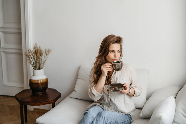 Μια νεαρή ξανθιά γυναίκα σε casual άνετα ρούχα κάθεται σε ένα λευκό καναπέ στο σπίτι και πίνει καφέ. Σκανδιναβικό εσωτερικό, μινιμαλισμός - Φωτογραφία, εικόνα