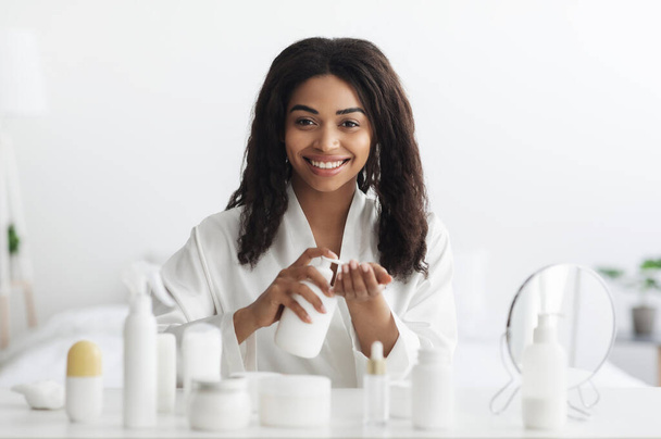 Cura della pelle quotidiana. Bella donna africana americana che pulisce il viso con latte detergente, seduta a tavola - Foto, immagini