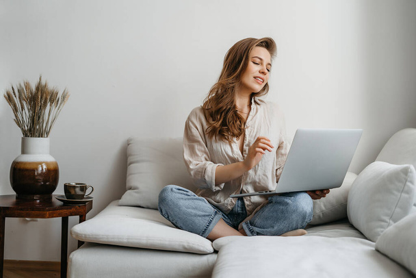 Happy smiling woman sitting on couch and using laptop at living room at home, watching funny video, learning language, video calling, mother working online - Photo, Image