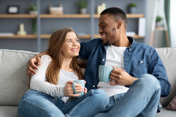 Занятия на выходных. Счастливая смешанная пара расслабляется на диване с кофе дома - Фото, изображение