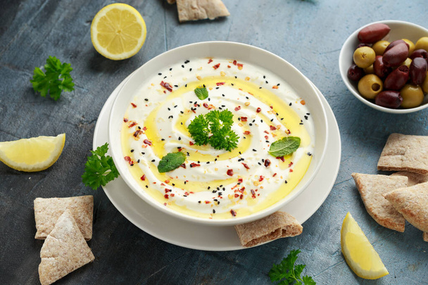Labneh Frischkäse-Dip mit Olivenöl, Salz, Kräutern serviert mit Oliven, Fladenbrot in weißer Schüssel - Foto, Bild