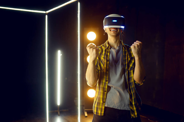 Hombre feliz usando auriculares de realidad virtual y gamepad en cubo luminoso, vista frontal. Interior oscuro del club de juego, foco en el fondo, tecnología VR con visión 3D - Foto, Imagen