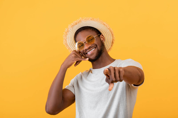 Chico negro alegre en traje de verano de moda que muestra llámame gesto, sonriendo a la cámara en el fondo del estudio amarillo - Foto, Imagen
