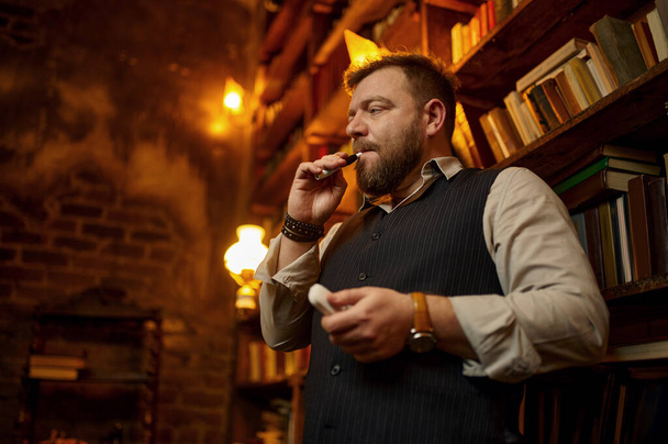 Γενειοφόρος άνθρωπος καπνίζει τσιγάρο με επιστόμιο, βιβλιοθήκη και πλούσιο εσωτερικό γραφείο στο παρασκήνιο. Καλλιέργεια καπνού, συγκεκριμένη γεύση - Φωτογραφία, εικόνα