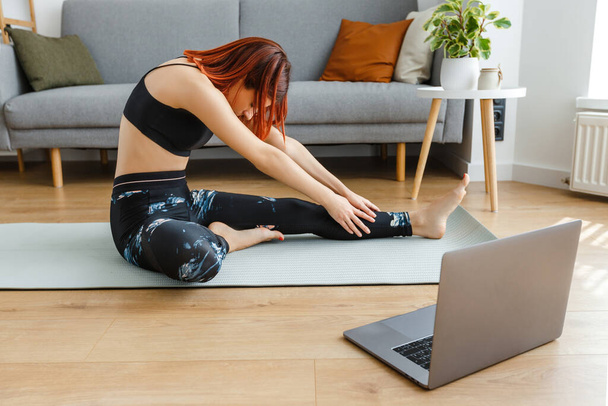 Junge, fitte Frau mit Laptop, die sich Online-Videostunden ansieht und zu Hause Yoga praktiziert. Fitnessübungen zum Dehnen. Gesunder und sportlicher Lebensstil. - Foto, Bild