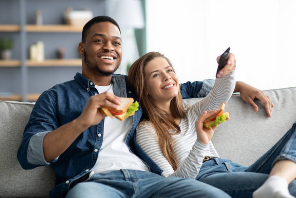 Weekend Relax. Cheerful Interracial Couple Eating Sandwiches And Watching Tv At Home - Photo, Image