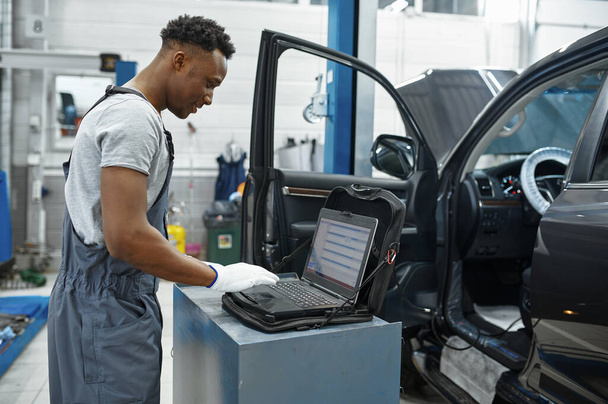 Male mechanic using laptop at the auto with opened hood, car service. Vehicle repairing garage, man in uniform, automobile station interior on background - Φωτογραφία, εικόνα