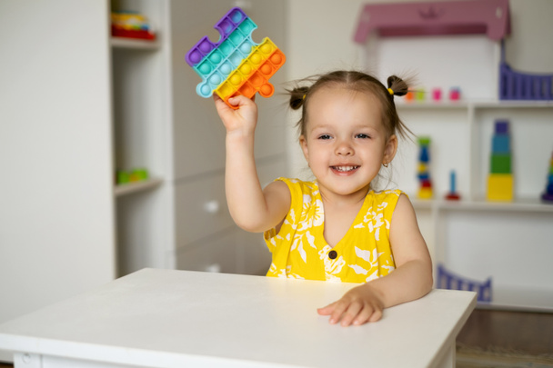 小さな女の子がおもちゃでテーブルで遊んでいます。開発のための魅惑的な感覚玩具。カラフルなケシのおもちゃ。シンプルなディンプル。その少女はおもちゃに情熱を注いでいる。笑いと楽しさ. - 写真・画像