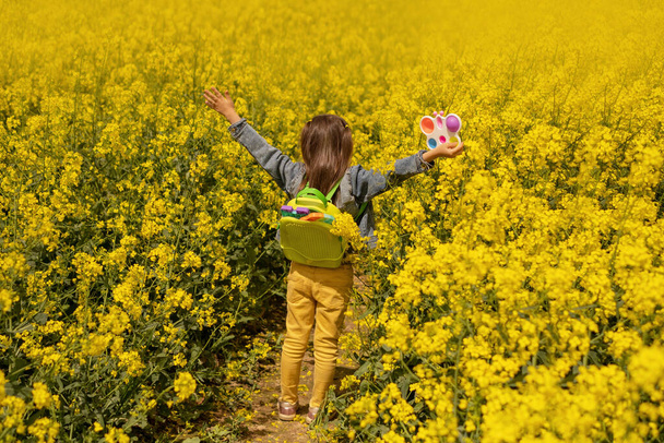Una chica en un campo de colza con una mochila llena de popit educativo moderno y simples juguetes hoyuelos. Un campo amarillo brillante de canola floreciente. Día soleado. Con los brazos abiertos. de pie con la espalda - Foto, imagen