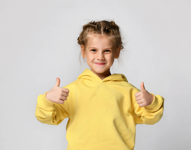 Portrait of a pretty attractive little girl in a yellow warm hoodie showing thumbs up with two hands on a white background. - Foto, Imagen