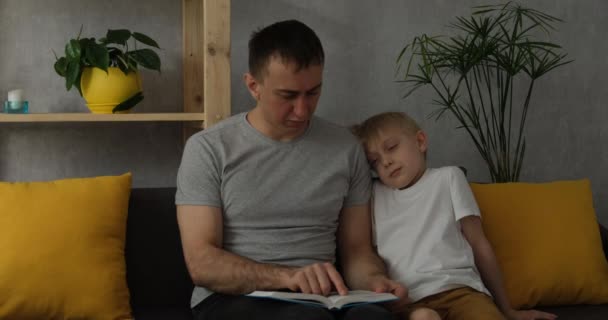 Papá le lee un libro a un niño dormido. El chico se durmió en el hombro de sus padres escuchando el libro. Padre e hijo leen sentados en el sofá - Metraje, vídeo