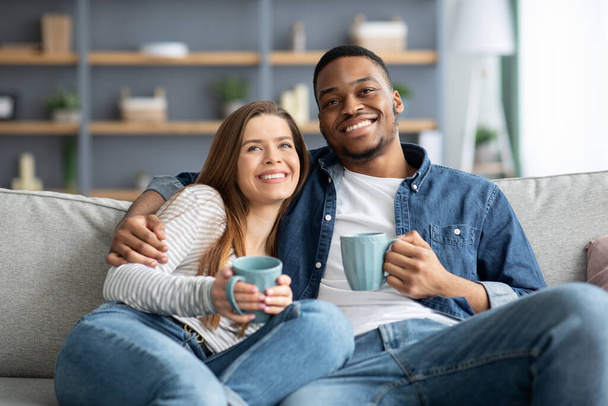 Σπίτι Αναψυχή. Χαρούμενο διαφυλετικό ζευγάρι βλέποντας τηλεόραση και πίνοντας καφέ στον καναπέ - Φωτογραφία, εικόνα
