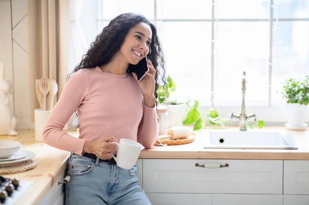 携帯電話で話す陽気なブルネットの女性とキッチンでコーヒーを飲む - 写真・画像