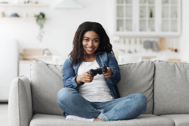 Amusant rester à la maison passe temps. Joyeuse femme afro-américaine utilisant un joystick pour jouer à des jeux vidéo en ligne, assise sur un canapé - Photo, image