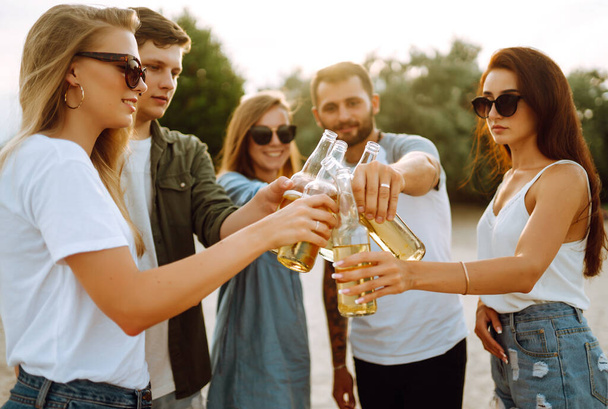 Group of friends cheers and drink beers on the beach. Young friends relaxing on the beath having picnic, toasting with beerus. People, lifestyle, travel, nature and vacations concept. - Foto, Bild
