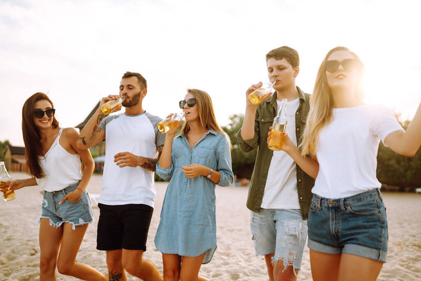 Group of friends cheers and drink beers on the beach. Young friends relaxing on the beath having picnic, toasting with beerus. People, lifestyle, travel, nature and vacations concept. - Foto, afbeelding