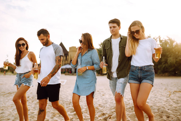 Group of friends cheers and drink beers on the beach. Young friends relaxing on the beath having picnic, toasting with beerus. People, lifestyle, travel, nature and vacations concept. - Фото, изображение
