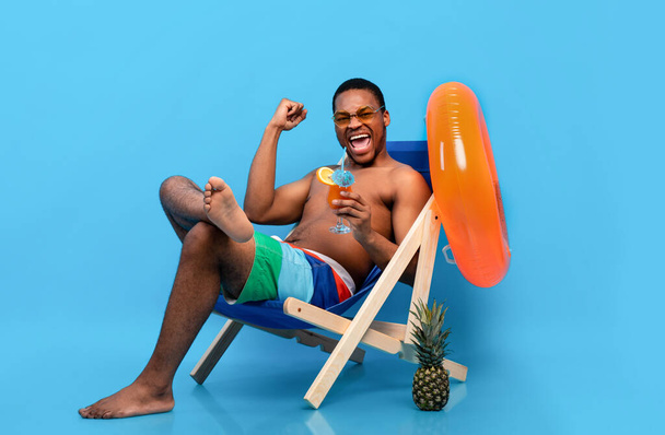Aufgeregter schwarzer Mann trinkt tropischen Cocktail im Liegestuhl und gestikuliert JA vor blauem Hintergrund - Foto, Bild