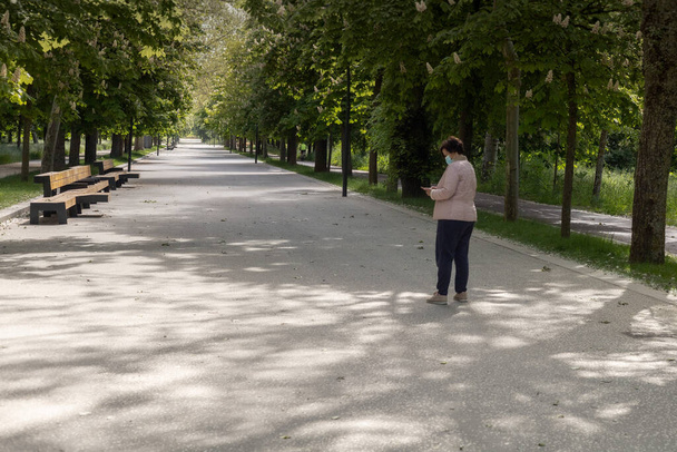 Женщина средних лет в коронавирусной маске пишет смс на смартфоне, прогуливаясь по парку, покрытому деревьями, с большой дорожкой в розовой куртке в солнечный день.. - Фото, изображение
