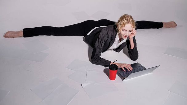 A ballerina dressed in a business suit poses for a laptop and drinks coffee. Flexible woman works at the computer - Foto, Imagen