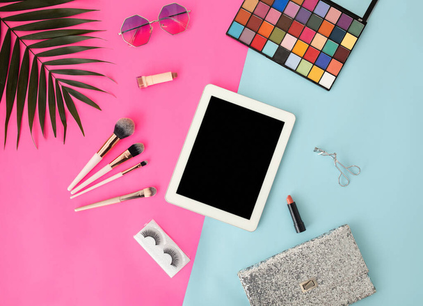 Tabletka z czystym ekranem i różnymi produktami do makijażu dla kobiet na niebieskim i różowym tle, widok z góry - Zdjęcie, obraz