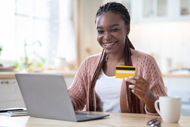Онлайн-платежи. Черная женщина держит кредитную карту и с помощью ноутбука на кухне - Фото, изображение