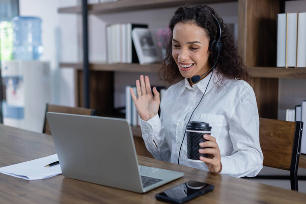 Mulher bonita usando fone de ouvido sentado na mesa movimento mão ter conversa on-line com laptop segurando xícara de café em casa escritório. - Foto, Imagem